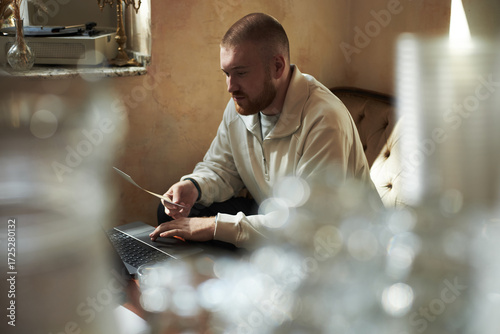 Fotografie Caucasian young adult man sitting on sofa working on laptop while holding docume