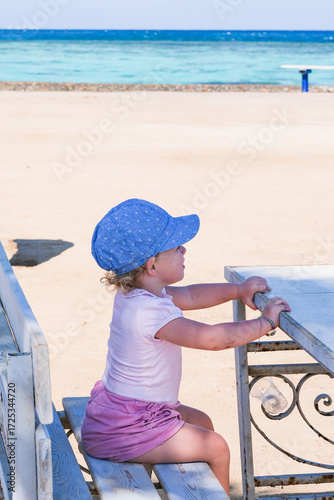 A young girl is sitting on a bench at the beach, wearing a blue hat