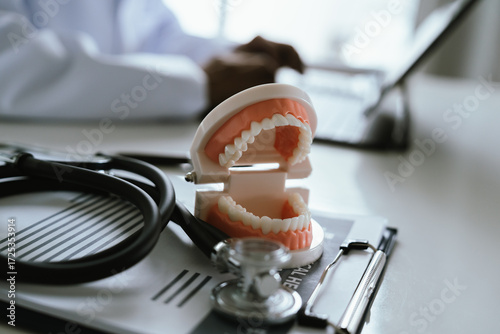 Doctor in white coat holding clipboard with medical documents, working at desk with laptop and dental model for healthcare planning.