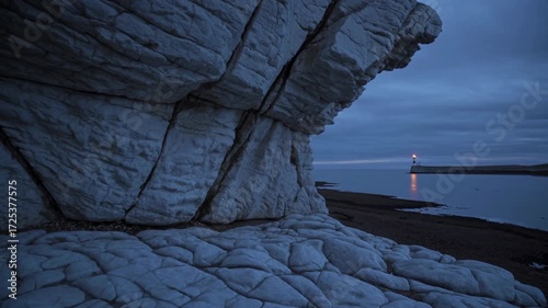 Dramatic View of a Chalk Cliff by the Sea with a Distant Lighthouse Illuminated at Dusk, Showcasing Tranquil Coastal Scenery Under Overcast Skies