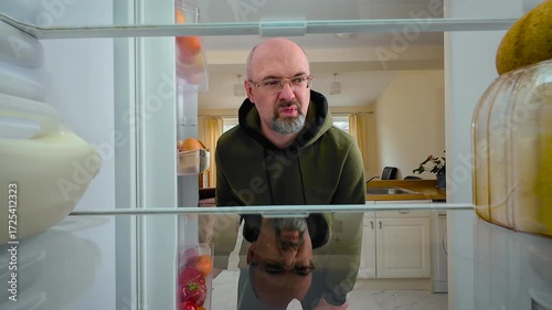 Man opening fridge and crying in frustration while seeing no food, view from inside of fridge. Male overwhelmed by hunger and sadness while looking at empty shelves. Guy wiping tears with distressed