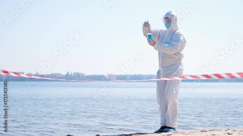 Person in protective hazmat suit and medical mask conducting water testing near lake. Environmental pollution research, contamination control, biohazard investigation, and safety inspection outdoors.