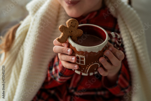 Closeup Little girl in pajamas sitting by the window drinking hot cocoa from a Christmas cup with a gingerbread man. Winter holidays.