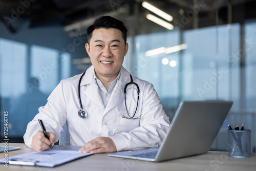 Close-up portrait of a young Asian male doctor in a white coat sitting at a desk with a laptop, making notes in a notebook