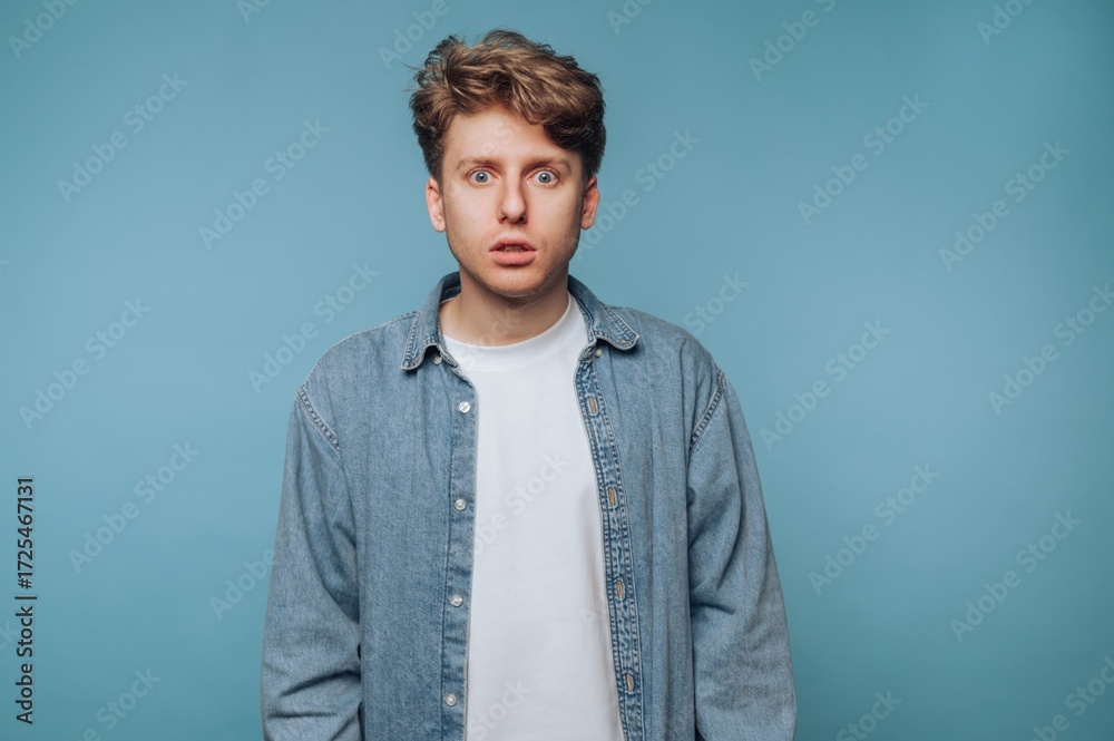 Obraz premium Young man in denim shirt looking surprised against blue background.