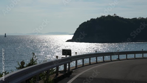 On Naoshima's winding coastal road, the Seto Inland Sea sparkles in the setting sun beyond guardrails, with lush islands and hazy mountains outlining tranquil summer coastal scenery.