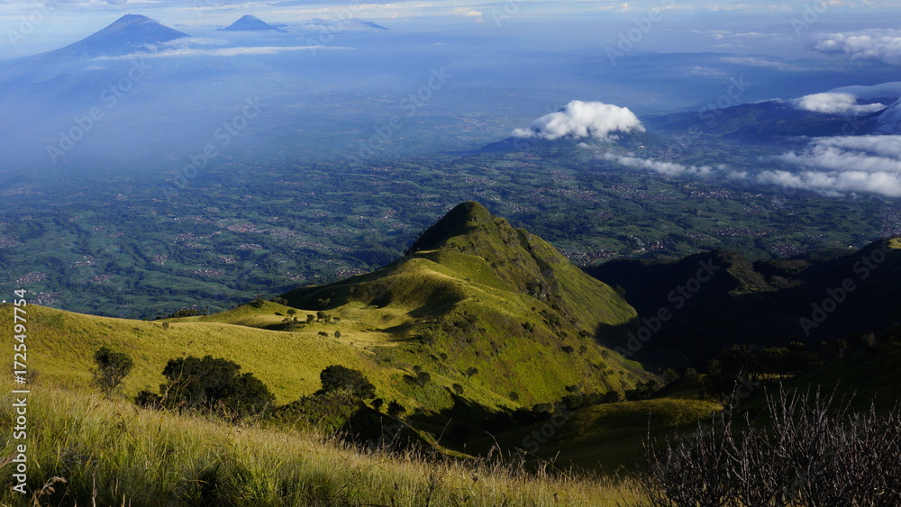 Fototapeta premium Expansive aerial view from a grassy mountain slope overlooking a vast valley with distant volcanoes piercing through the clouds.