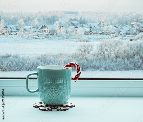 Winter background, cup with candy cane on windowsill and winter rural landscape outdoors. Cozy still life with concept of spending winter Christmas time indoors
