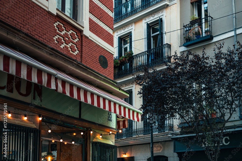 Obraz premium A charming building in Madrid featuring striped awnings, iron balconies, and warm evening lights in a lively neighborhood.
