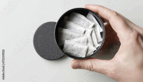 A man holds a round black container filled with neatly packed snus packets intended for oral use. The setting is neutral with a simple background, emphasizing the product