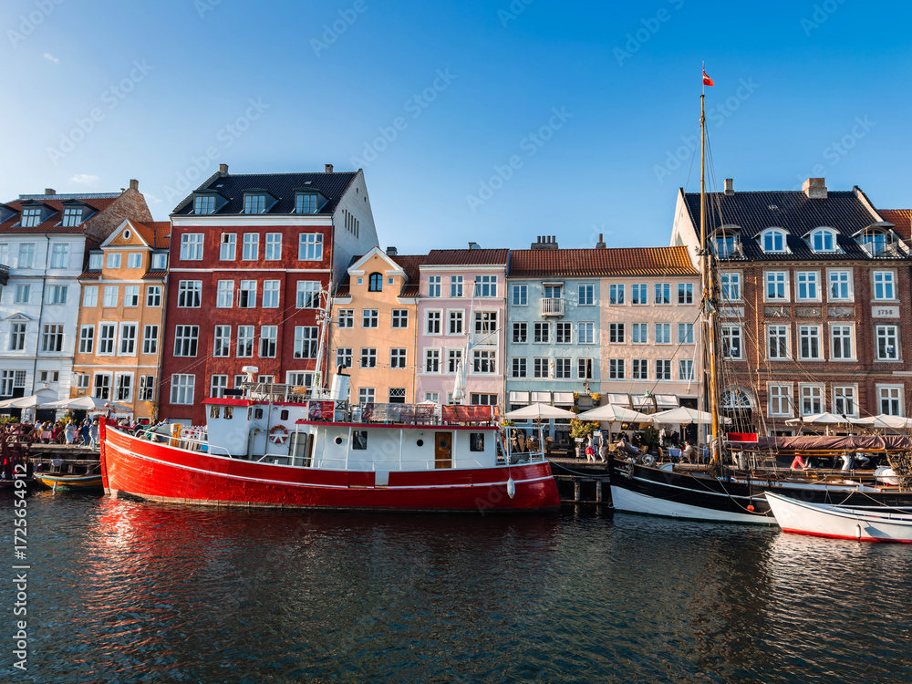 Fototapeta premium The Nyhavn district in Copenhagen, Denmark, features colorful historic buildings, a red boat in the canal, smaller vessels, and outdoor cafes under a clear sky.