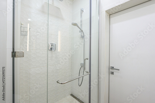 A modern bathroom features a glass-enclosed shower with white textured tiles. Chrome fixtures and a white door provide a clean, minimalist aesthetic