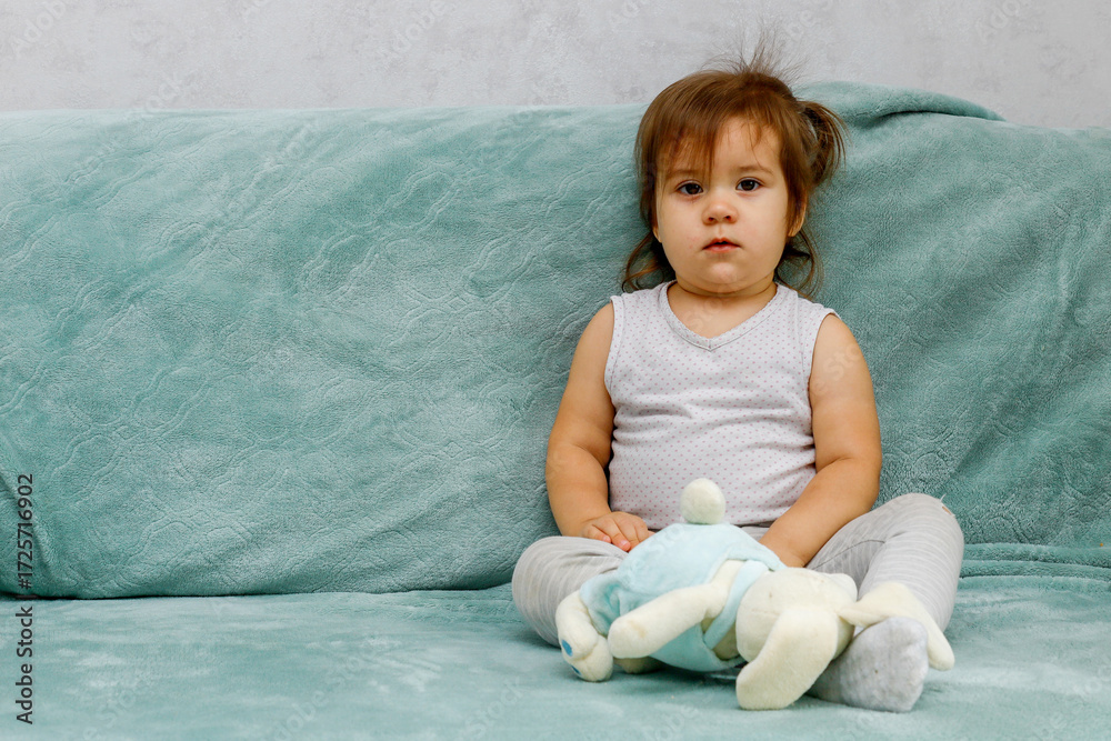 Fototapeta premium A little girl sits on a sofa with a toy in her hands and looks attentively into the camera