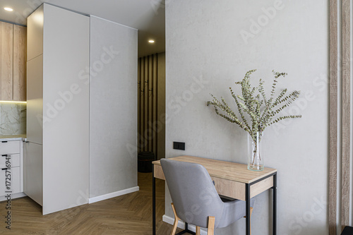 A modern interior shows a desk with a vase of plants, a gray chair, and a glimpse into a hallway with wooden accents