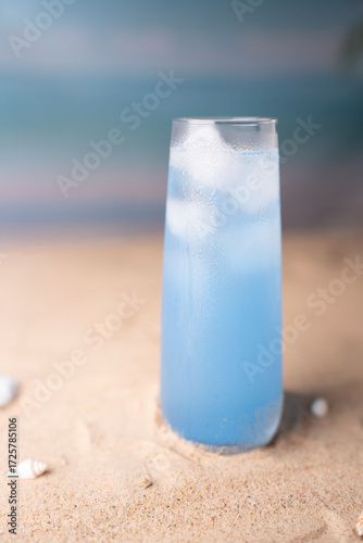 refreshing blue drink in frosted glass with ice cubes on sandy beach with subtle sea view evoking a relaxing tropical summer ambiance, rejuvenate, nature, peaceful, bliss, tranquility, waves