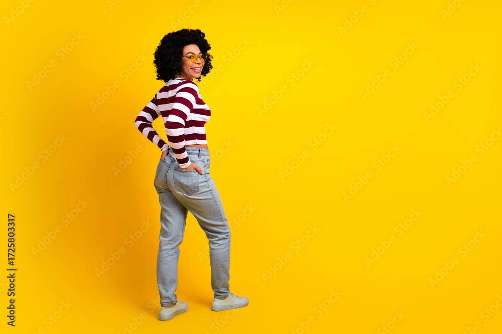 Fototapeta premium Young stylish woman with curly hair wearing striped pullover and jeans posing confidently against bright yellow background
