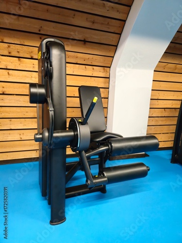 A sleek black leg extension and curl machine stands on a blue gym floor against a modern wooden wall.