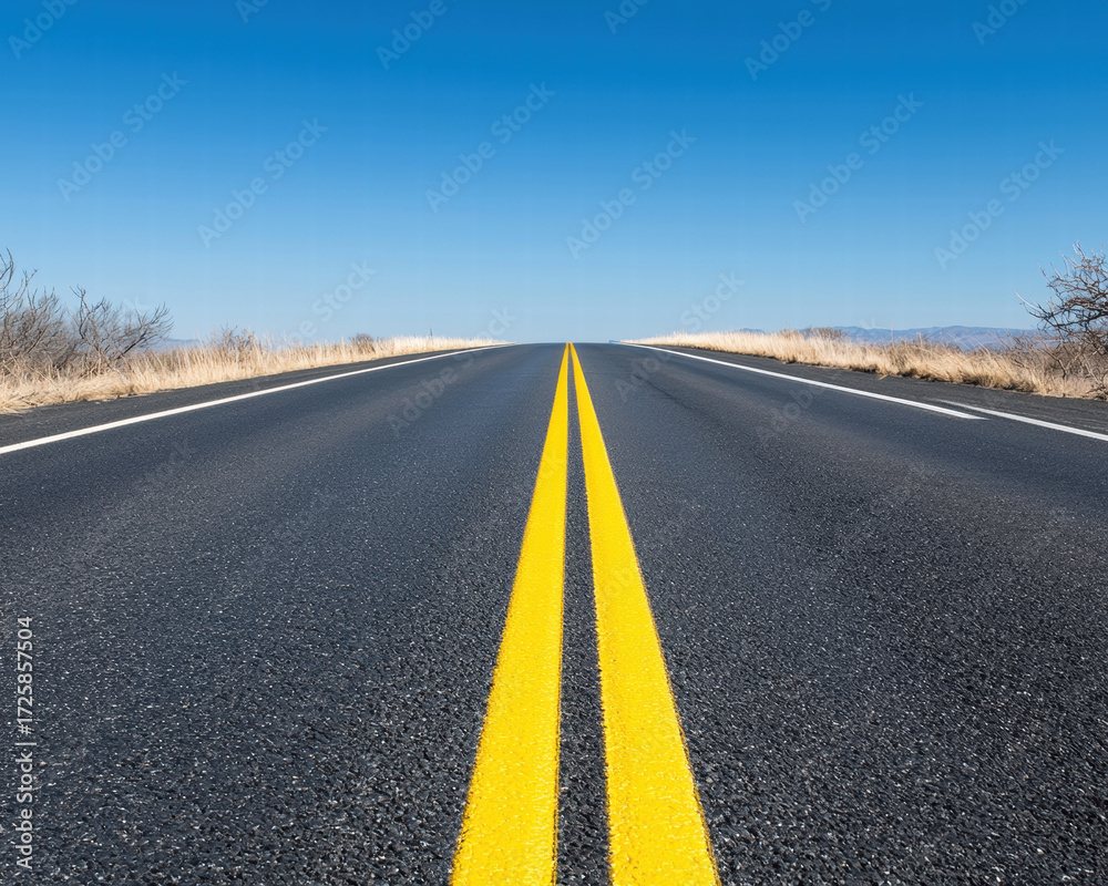Naklejka premium Lonely road stretching into horizon with vivid yellow centerline under clear blue sky evoking solitude and journey
