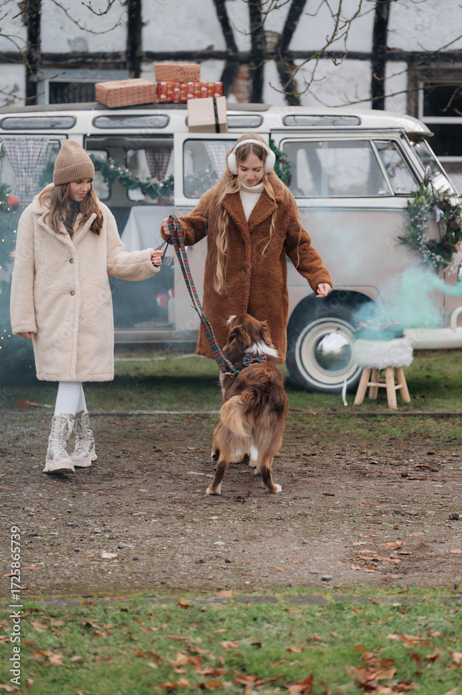 Obraz premium Two women in cozy winter clothes with a dog in front of a decorated camper van, enjoying festive outdoor holidays with gifts and seasonal decorations. 