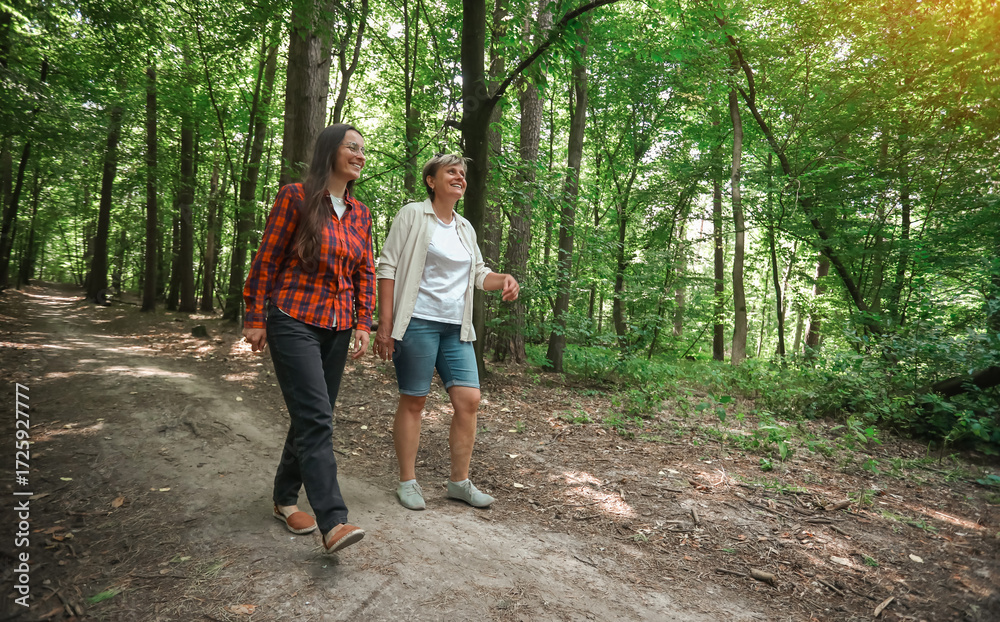Fototapeta premium Two women walking together in a green forest, smiling and enjoying nature on a summer day. Friendship, leisure, outdoor activity, and healthy lifestyle.