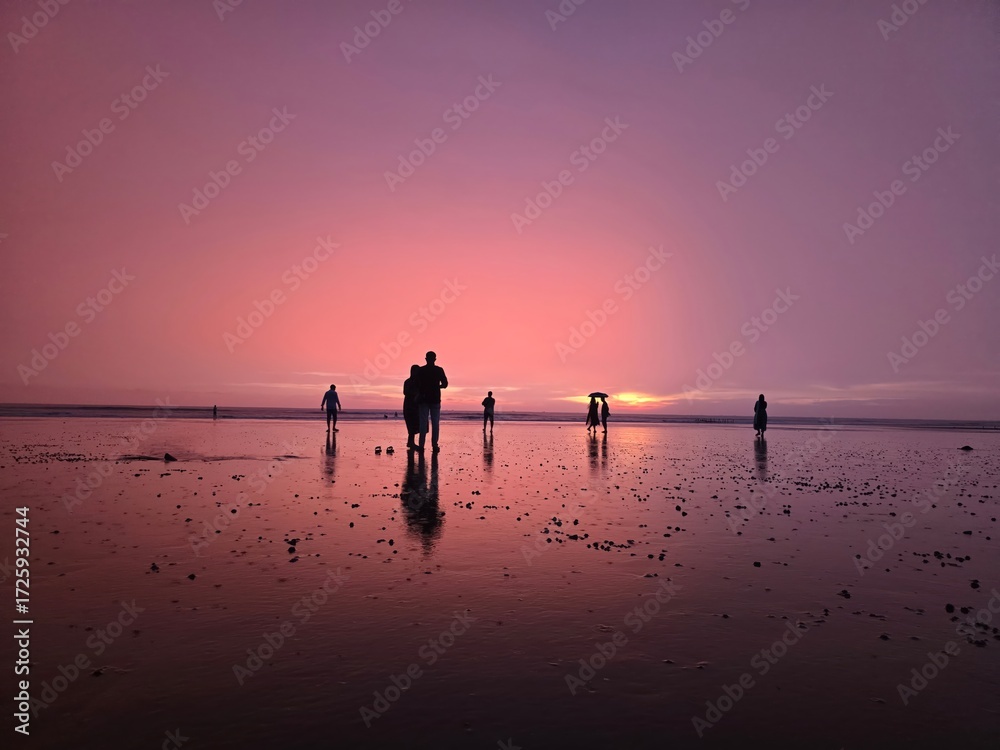 Fototapeta premium Silhouettes of people walking on a wet beach during a vibrant pink and purple sunset, reflecting on the water