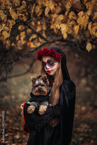 A young woman in a Day of the Dead costume: purple and violet skull makeup, red roses on a wreath, and a black cloak. She held a small dog against the backdrop of an autumn park with fallen leaves.