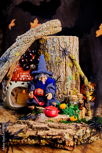 postcard with a halloween witch in a pointy hat with spiders, bottles, a saucepan and a ladle sits on a stump under a snag and brews a potion in a gloomy forest on All Saints Day, selective focus