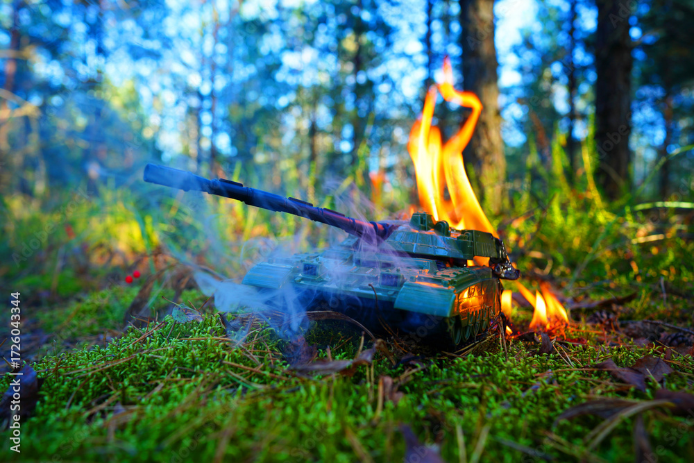 Naklejka premium Military Toy Tank Catches Fire in Forest During Bright Sunny. Smoke rises against of tall trees. Game of War. Battle tank during burning. War costs, military spending. Battle toy tank in burnt.