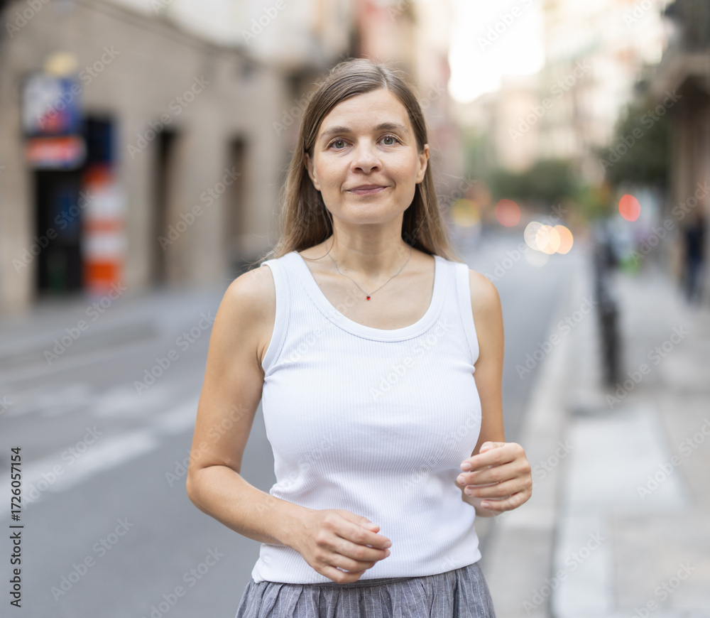 Fototapeta premium Positive woman walks along sunlit street, walks around city, spends time in public urban space. Person and megalopolis, solitude or loneliness .
