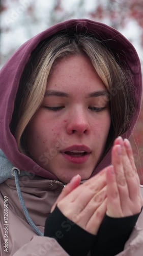 Woman in hooded winter jacket standing in cold, crossing arms to warm up, blurred background features trees with red berries, creating a serene winter atmosphere