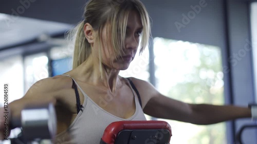 Shoulder Workout: Strong blonde woman doing lateral dumbbell raises on a bench in the fitness club