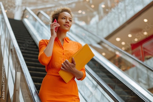 Woman in orange dress holds phone and yellow file on escalator in building