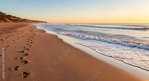 Empty beach at sunrise with gentle waves, footprints in sand leading to ocean, no people visible