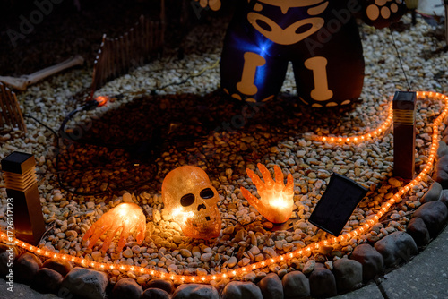 Glowing skull and hands sit on gravel with orange rope lights at night. Spooky Halloween yard decoration with stones and eerie mood.