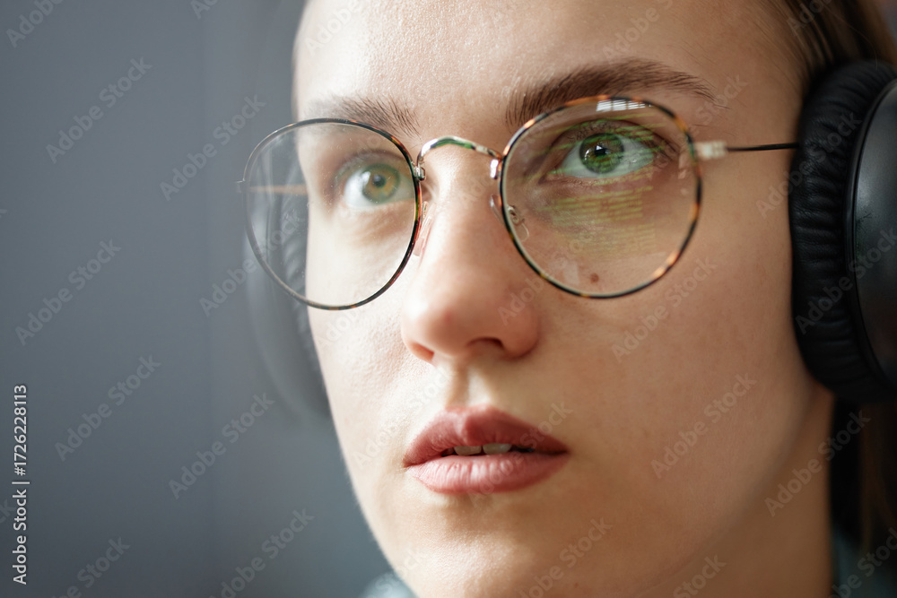 Fototapeta premium Young adult Caucasian woman wearing eyeglasses and headphones looking at computer screen with code reflected in lenses showing focused facial expression