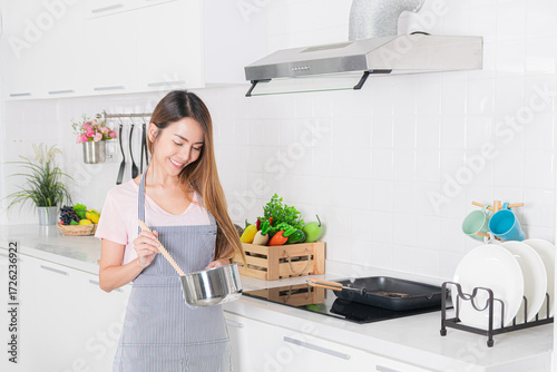 Smiling asian woman in a striped apron hold a pot and wooden spoon ready to cook in her bright clean and well-equipped modern kitchen.