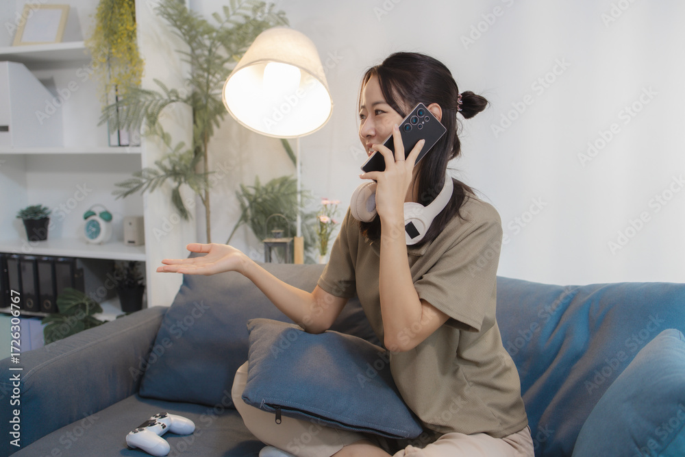 Naklejka premium A cheerful young woman sits casually on a sofa, holding a smartphone to her ear while talking and smiling. With headphones around her neck, she looks relaxed, expressive, and engaged.