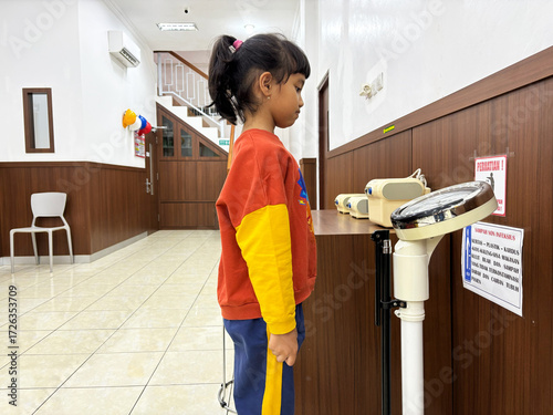 Photos Little girl weighing herself on scale at doctor's office during routine checkup, showing focus and determination to monitor her health and growth