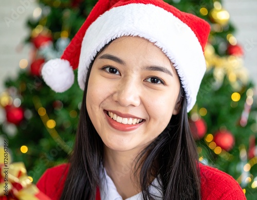 A woman in a red Santa hat stands near a festive Christmas tree, her hair dark and straight, with blurred ornaments and golden lights in the background