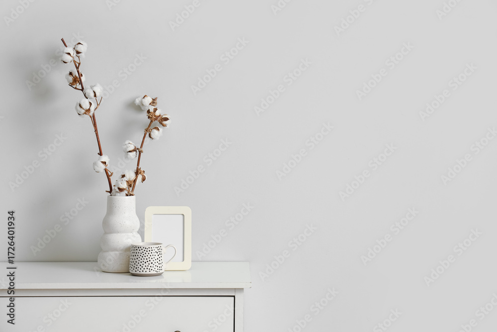 Fototapeta premium Photo frame, cup and vase with cotton flowers on chest of drawers near white wall in living room, closeup