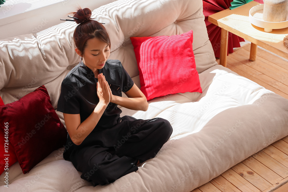 Obraz premium Young asian woman meditating in prayer pose on sofa, practicing mindfulness and relaxation at home, she is sitting cross-legged with her eyes closed and hands folded