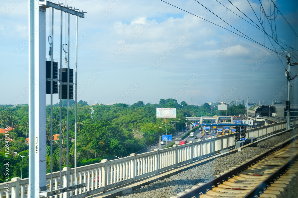 Fototapeta premium View from the Jakarta–Bandung high-speed train window showing elevated railway tracks above a busy toll road with green trees and a bright blue sky in the background.