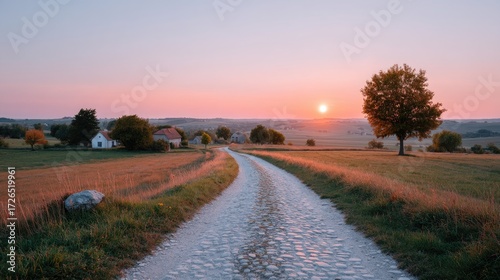 Cobblestone Country Road Through Rural Meadow Landscape at Sunset with Pink Sky and Distant Houses