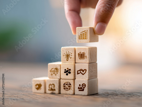 Hand stacking wooden block cubes with engraved business icons like growth charts, lightbulb, and globe, symbolizing strategic planning, step-by-step success, corporate development, and foundation buil
