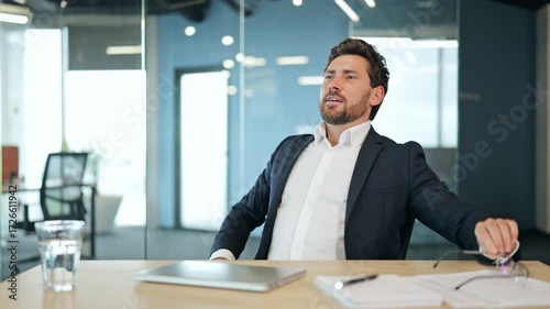 Satisfied professional businessman in a dark suit and white shirt at an office desk closes laptop, removes glasses, and leans back, feeling happy and content in a modern office.