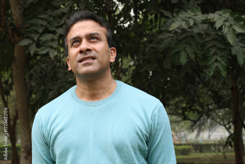 Portrait of a smiling young Indian man standing in casual tshirt in a park, looking confidently at the camera. resting, relaxation feel satisfied glad expressions