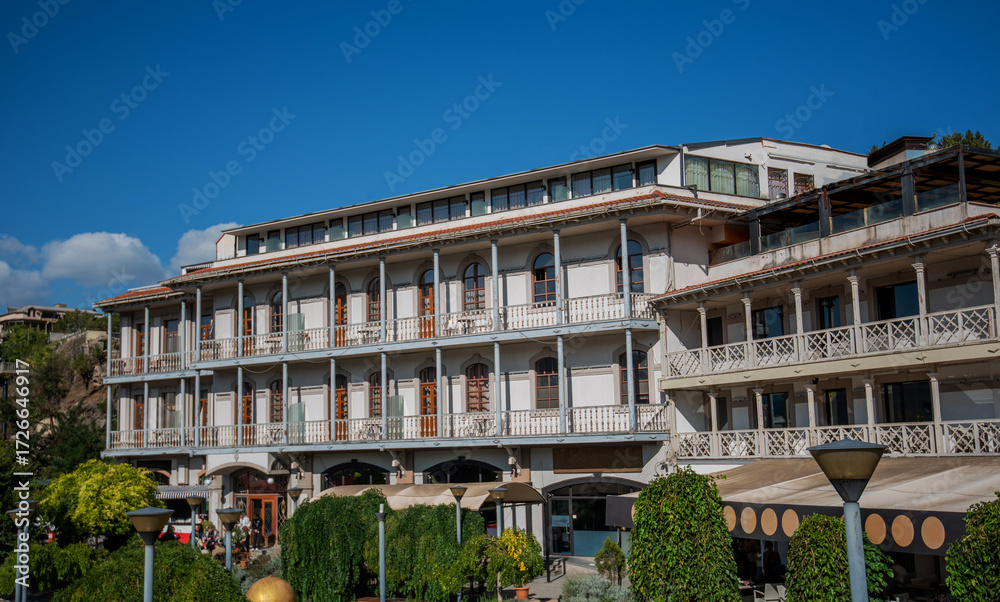 Fototapeta premium A traditional Georgian building with arched windows and wooden balconies in Tbilisi. Surrounded by greenery and sunny weather, the structure features elegant architecture and historical charm.
