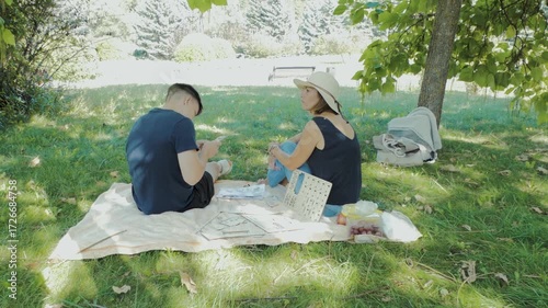 A couple enjoys a leisurely picnic in a sunny park, assembling a wooden puzzle or model together under the shade of a tree. Relaxation, hobby, and togetherness outdoors.