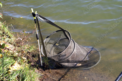 Fishing keepnet made of black mesh is fixed with strap and metal holder on shore, partly submerged in lake water, ready to store caught fish during angling. Fisherman equipment. Fisherman tools.