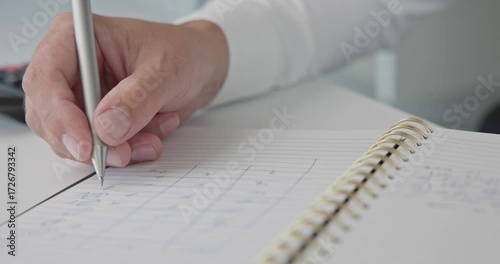 Close-up of hands - a man in a shirt draws and fills a table with a pen in a notepad on a metal spring on an office desk